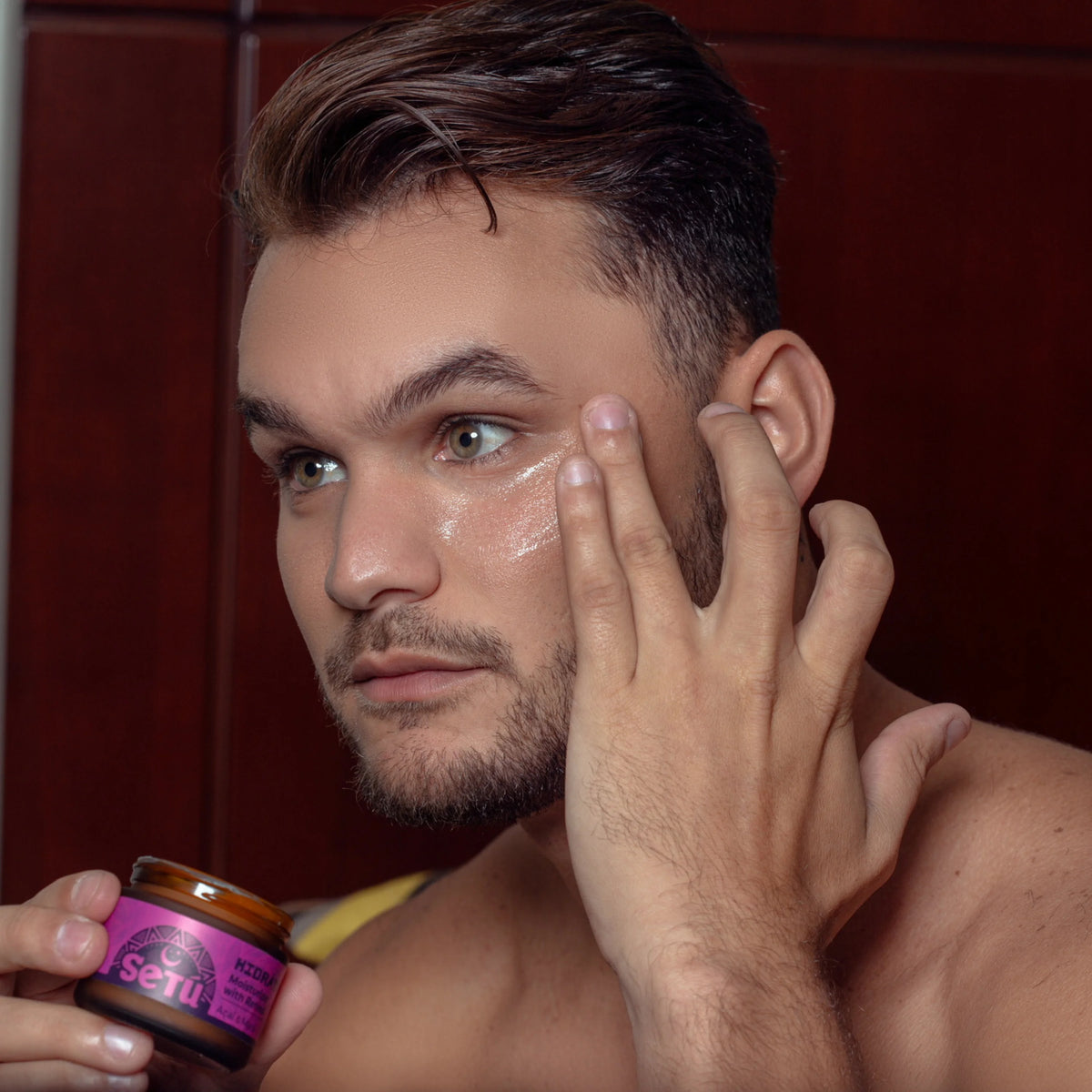 Man applying setu moisturizer with retinol, acai, and maca to his face with a jar of cream in hand, against a wooden background.