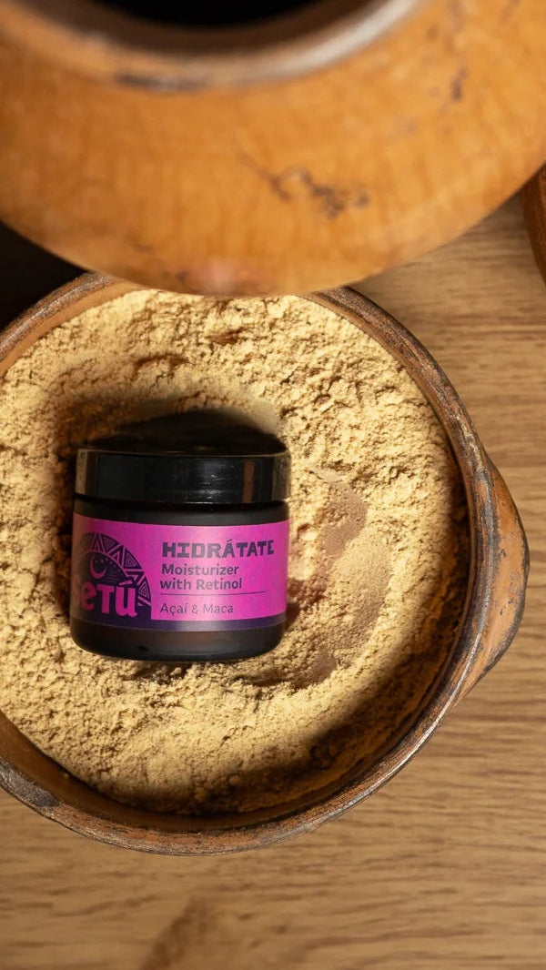 A small jar of maca root powder with a purple label of setu hidratate moisturizer for men sits in a wooden bowl filled with light tan powder, alongside a wooden spoon with more powder and a rustic wooden pitcher on a wood-grain surface.