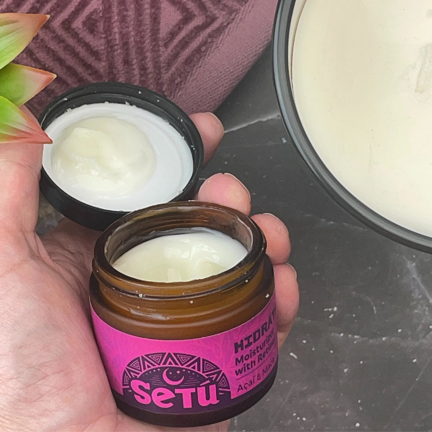 Hand holding a jar of setu moisturizer for men with retinol, acai, and maca with pink label, open lid showing cream, and blurred background.