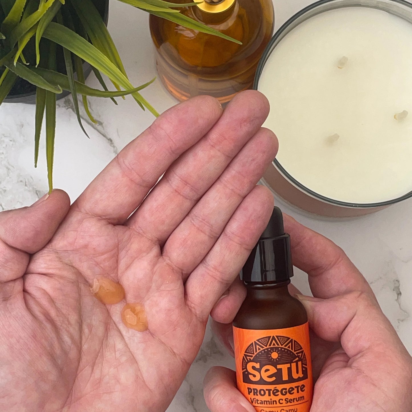 Person holding a bottle of 'Setu' skincare serum with a candle and plant in the background.