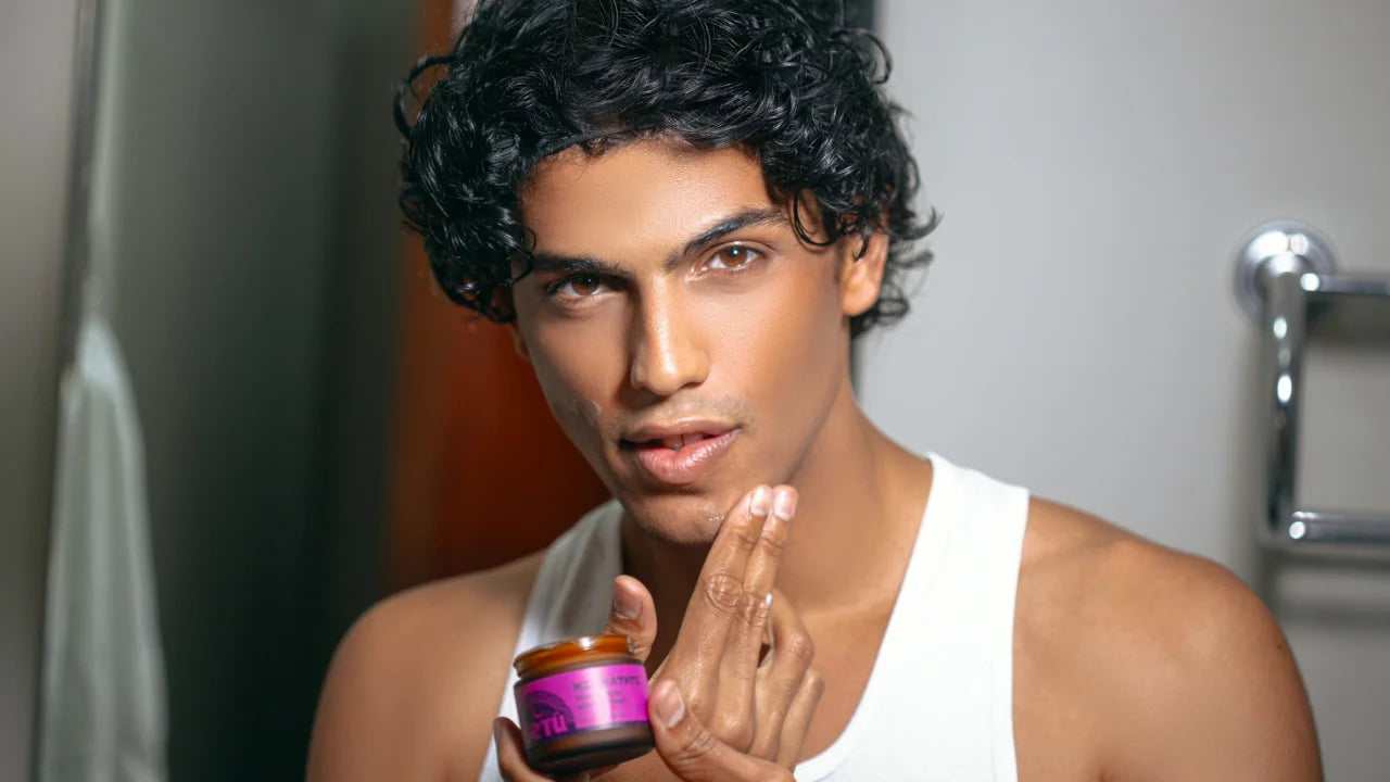 A man with curly dark hair wearing a white tank top holds a small jar with a pink label and amber lid, touching his face as if applying product. The background is a softly blurred bathroom setting with gray walls and a chrome fixture.