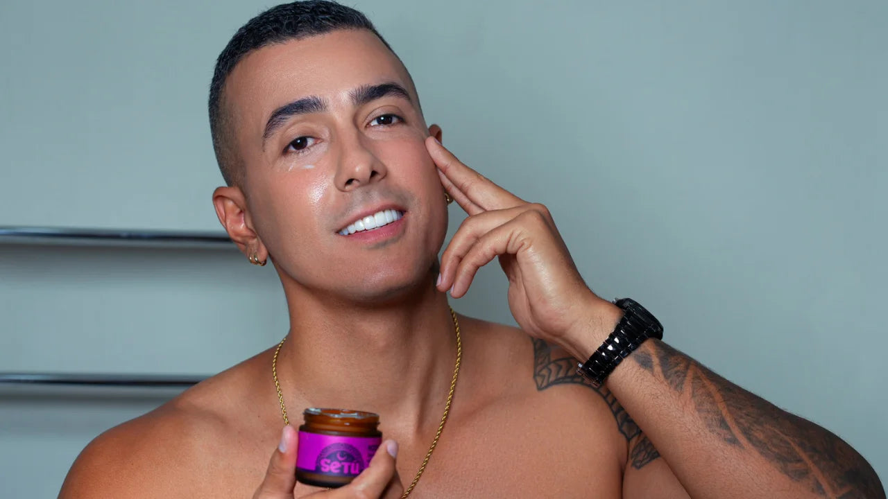 A shirtless man with tattoos and a gold chain holds a small jar of SETU natural skincare product while touching his face, against a muted blue-gray background.