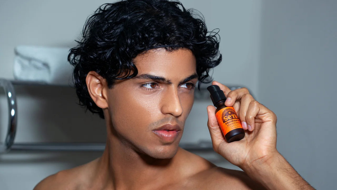 A man with curly dark hair holds a small orange and black bottle near his face in a bathroom setting.