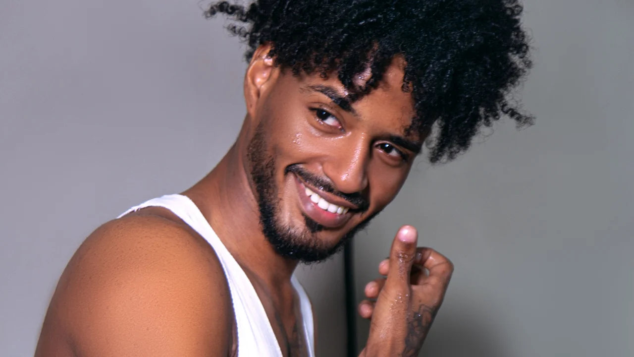 A smiling man with curly black hair and a beard, wearing a white tank top, posing against a solid gray background.