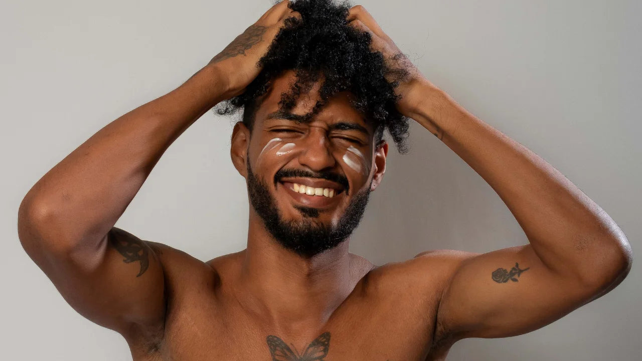 A shirtless man with curly black hair and tattoos poses with his hands in his hair, smiling broadly. He has eye cream on his cheeks and forehead, set against a plain light gray background.