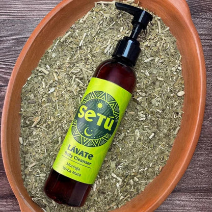 Setu Lavate daily cleanser bottle with green label in a terracotta pot filled with dried herbs.