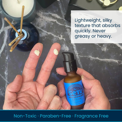 A person holds a small amber bottle labeled SETU Eye cream against their hand, with white eye drops visible on two fingers. The scene is photographed on a dark marble-like surface with a candle and reed diffuser in the background.
