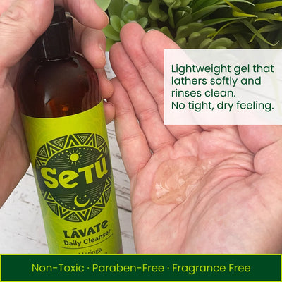 A person holds a bottle of Setu Lavate Daily Cleanser next to their open palm showing a clear gel. The bottle has a bright lime green label and an amber brown bottle.