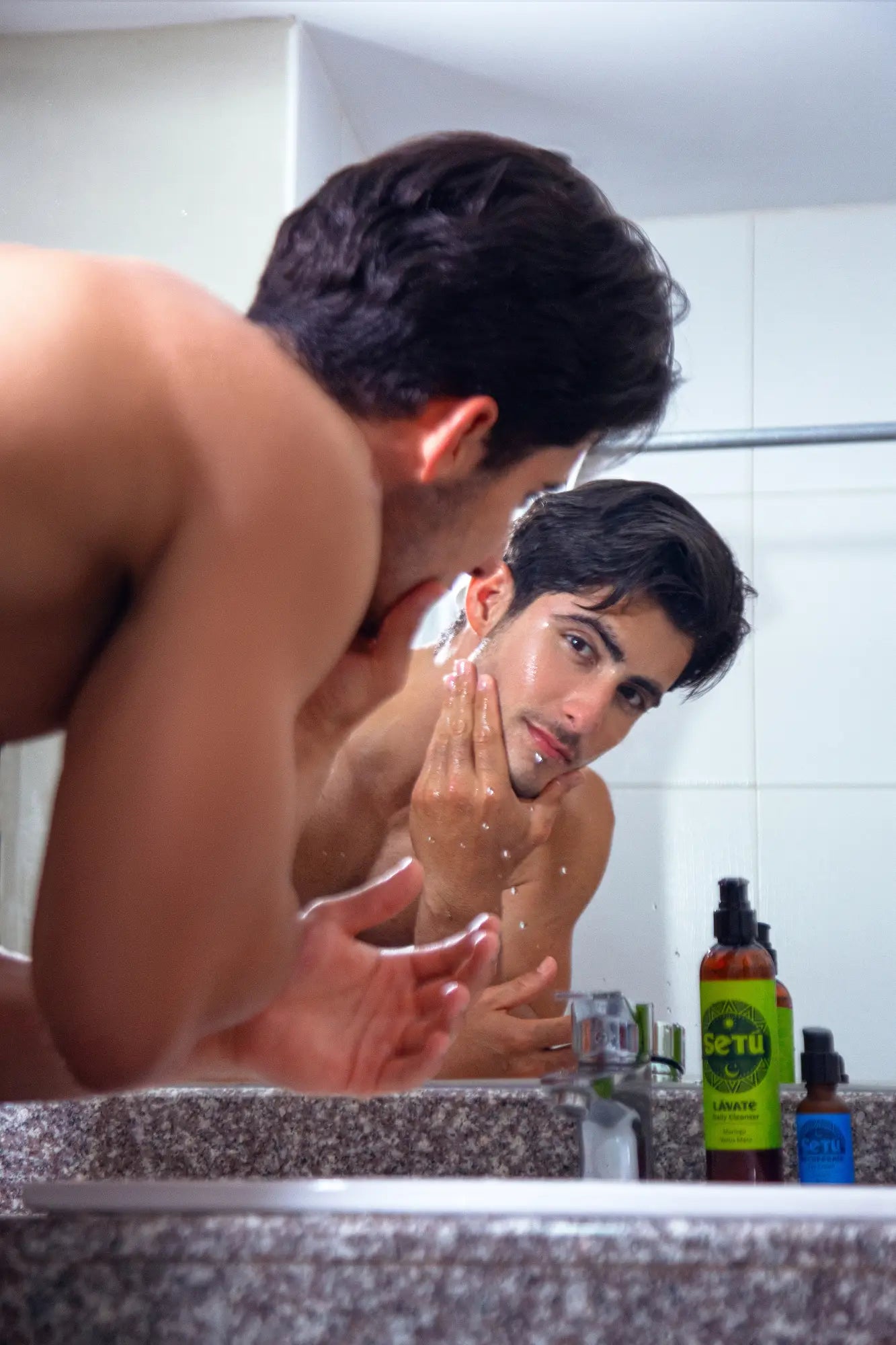 A shirtless man in a bathroom looks into a mirror while washing his face, with Setu bottles on the counter in the foreground.