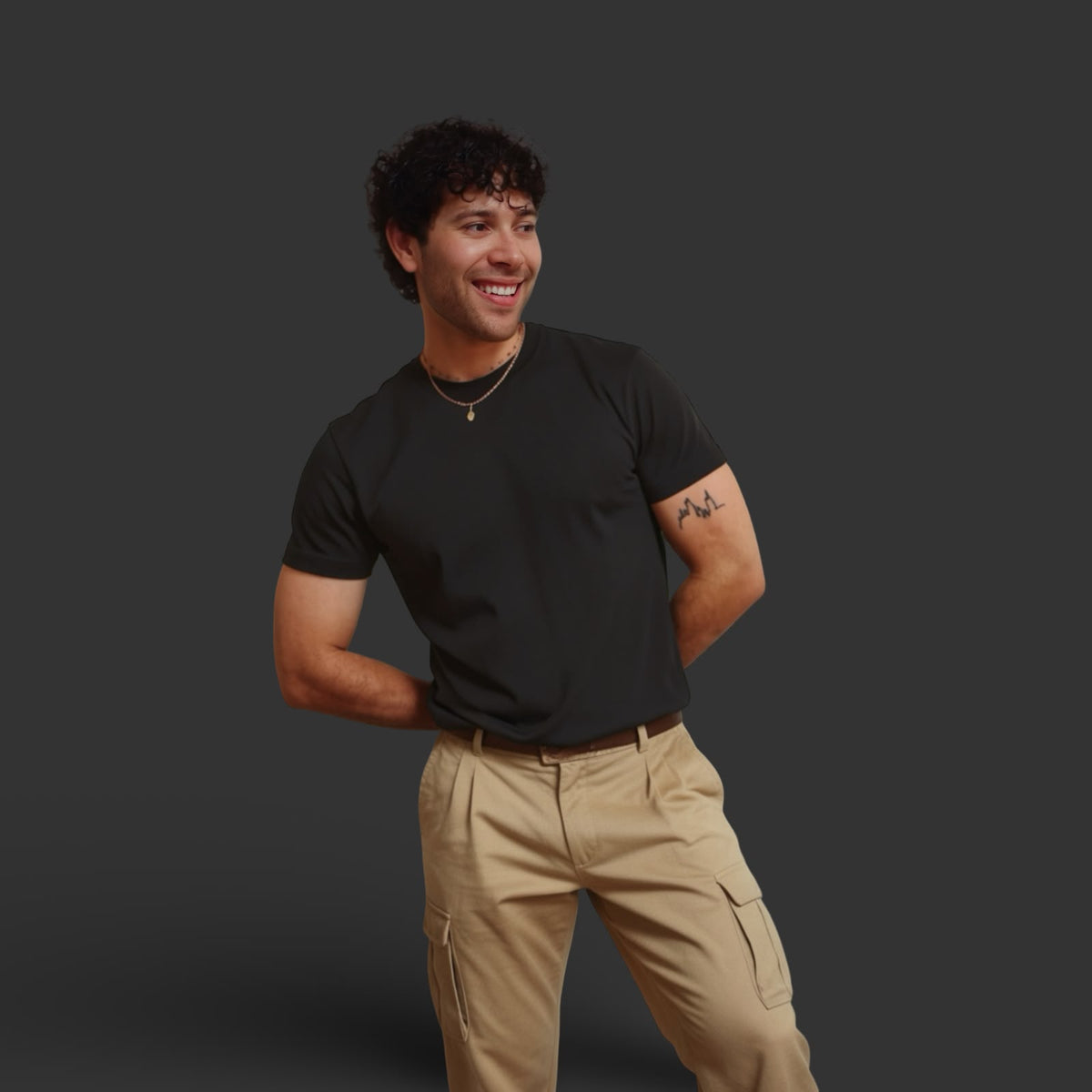 A man smiling and looking to the side, wearing a black crew-neck t-shirt and beige cargo pants, standing against a solid dark gray background.