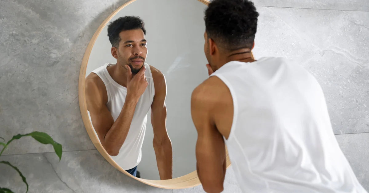 A man in a white tank top stands in front of a round wooden-framed mirror, looking at his reflection while shaving. The scene is set in a modern bathroom with a gray stone wall.