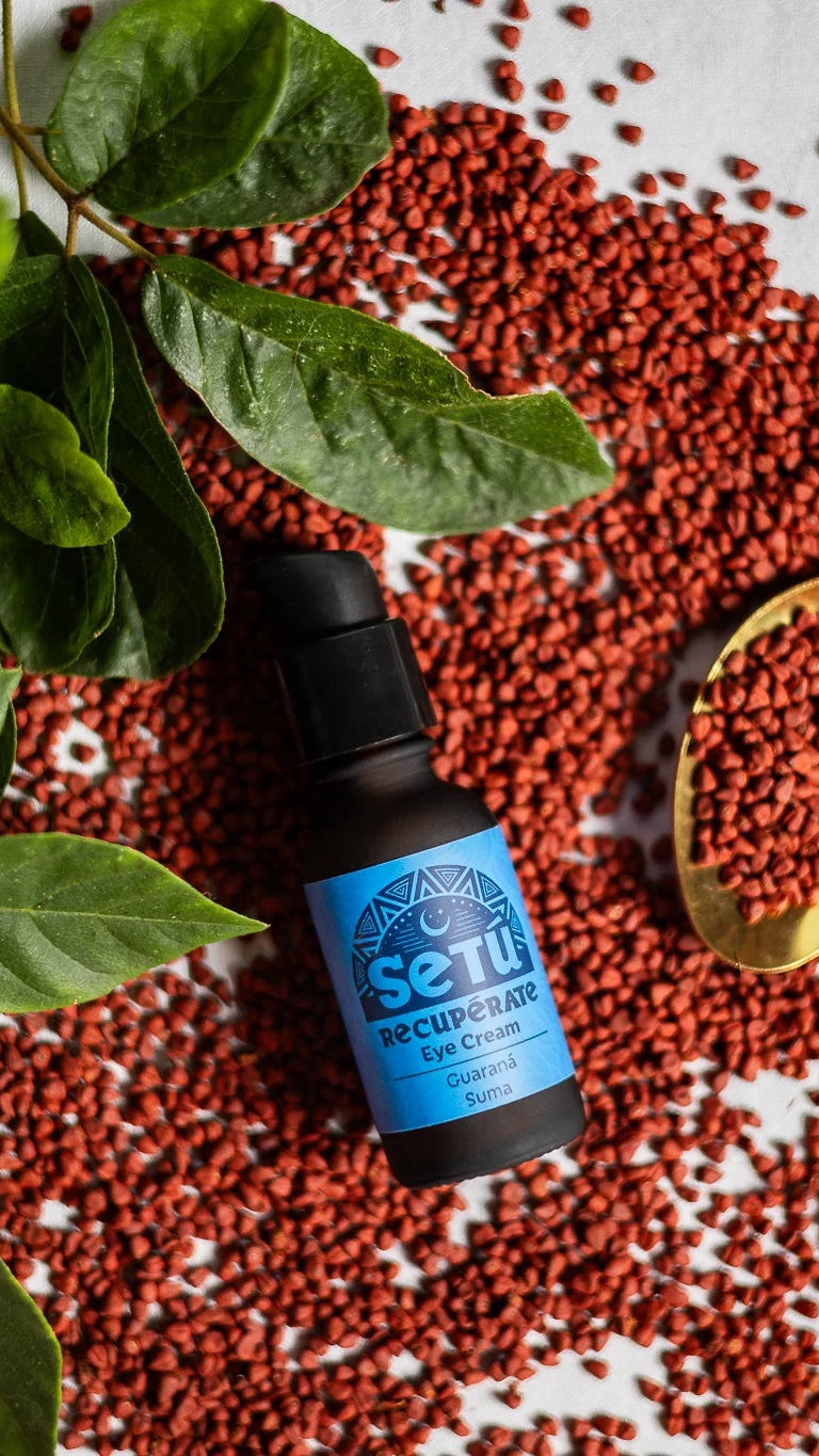 A small bottle of Setu Eye Cream for Men with Achiote with a blue label and black dropper cap, styled on a white surface with scattered red Achiote berries, green leaves, and a gold spoon.