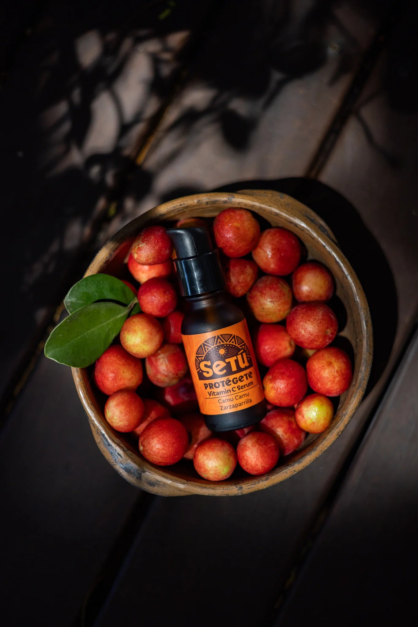 A small amber bottle labeled Setu protegete vitamin c serum rests on top of a bowl filled with camu camu red and yellow fruits, photographed from above on a dark wooden surface with leaf shadows.