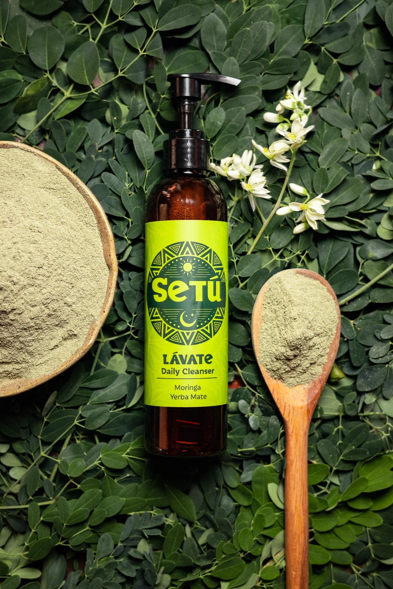 A bottle of Setú Lavate Daily Cleanser with a bright lime green label and black pump, styled on a bed of green leaves with a wooden spoon and bowl of powder.